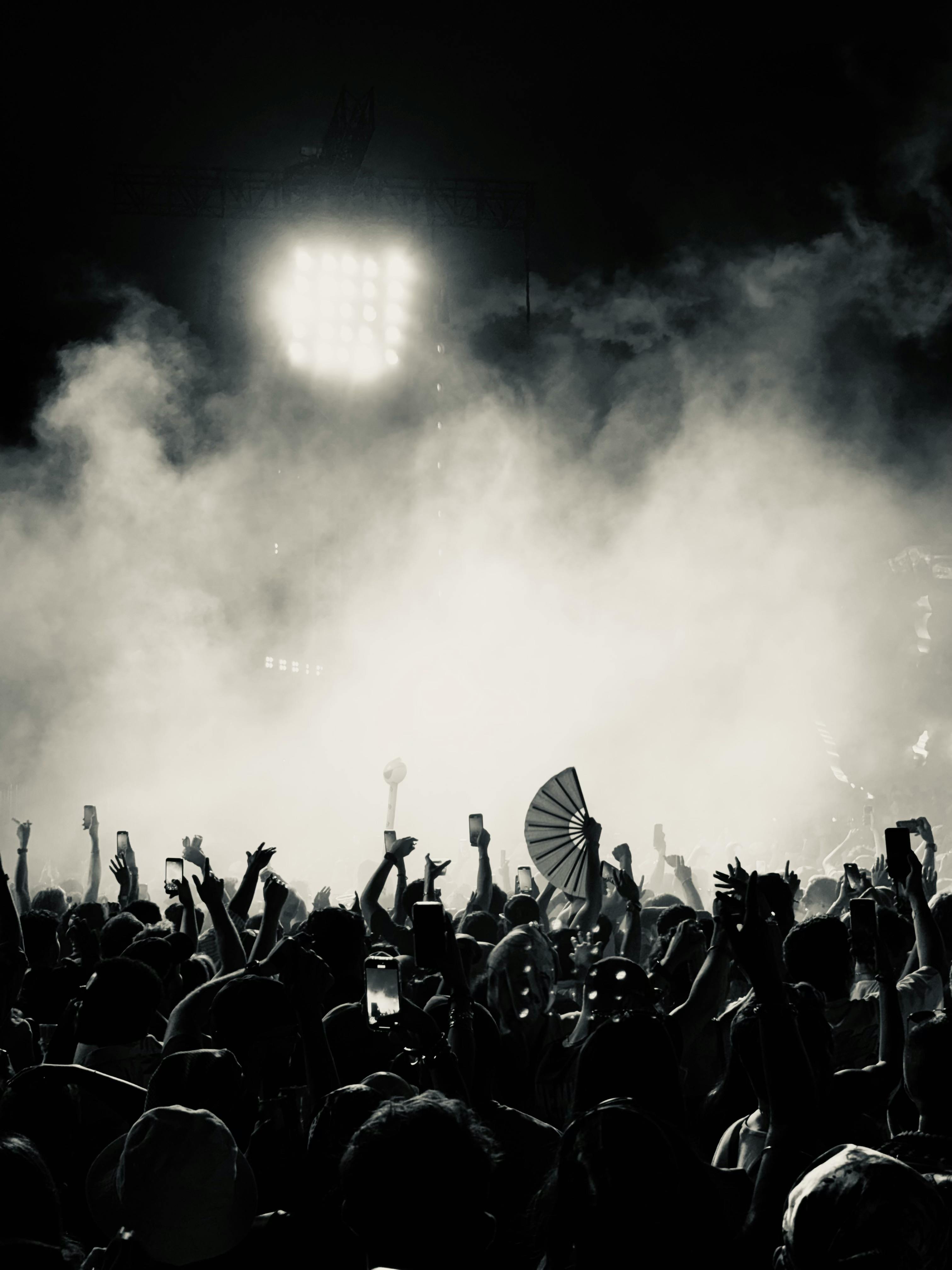 Dramatic black and white shot of festival crowd with hands raised and fan visible, stage lights cutting through smoke